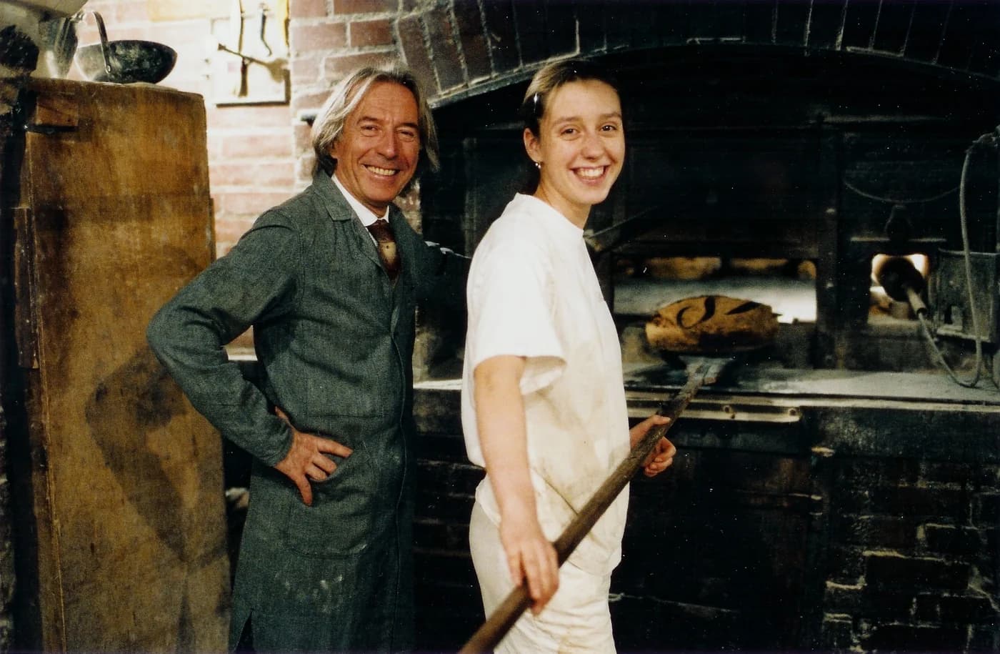 Two bakers smiling beside a traditional wood-fired oven.
